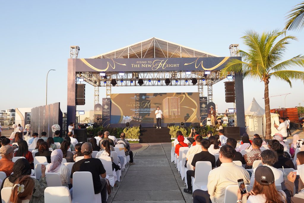 outdoor stage event with a decorated banner reading rising to the new height and an audience seated in white chairs under a clear sky   citraland city center point indonesia